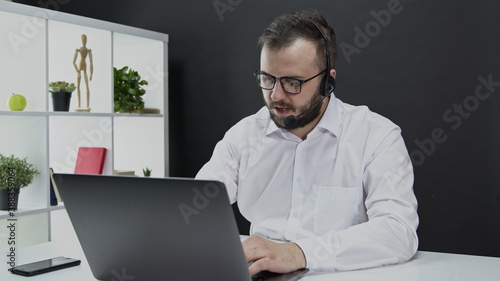 A technical support specialist advises the client. Caucasian bearded man in a white shirt in the office at the computer and in wireless headphones. Counseling people on the hotline. 24/7 user support.