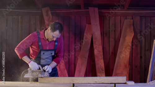 Male carpenter cleans the surface of a wooden beam using an electric planer in the workshop. Work with electric planer for polish boards and bar. Home furniture manufacturing concept.