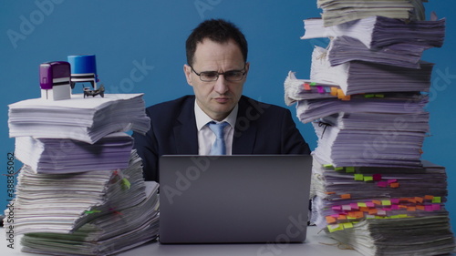 Concentrated office worker in shirt, tie and glasses sits at desk littered with papers, stamps on stack of documents. Serious manager working on laptop among piles of paperwork, overloaded with work