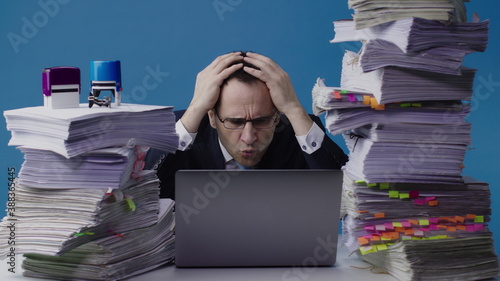 Concentrated office worker businessman working on laptop among piles of paperwork and stamps on stack of documents overloaded with work. Grabbing his head because of an mistake, deadline concept