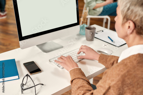 A mature ladyboss wearing fashionable clothes with a short haircut typing on a keyboard while sitting in front of a PC at a table photo