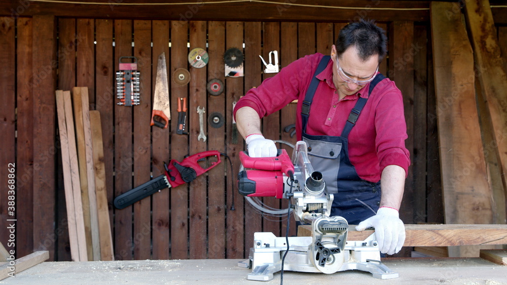 Male joiner processes wood bar using a miter saw in a workshop. Work with miter saw for cutting boards and bars. Home renovation with professional construction and repair tool. Male joiner processes wood bar using a miter saw in a workshop. Work with miter saw for cutting boards and bars. Home renovation with professional construction and repair tool.