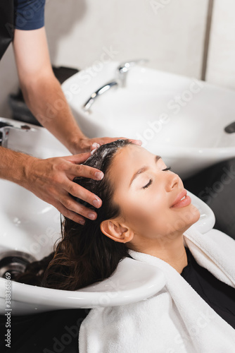 young woman smiling with closed eyes while barber washing her hair in beauty salon