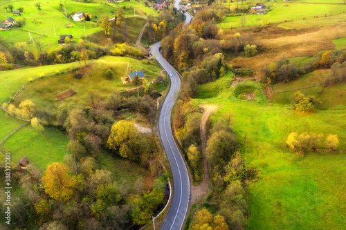 Aerial view of beautiful winding road in green hills at sunset in autumn. Colorful landscape with rural road, trees, grass and buildings in fall. Top view of curved roadway mountain village. Travel
