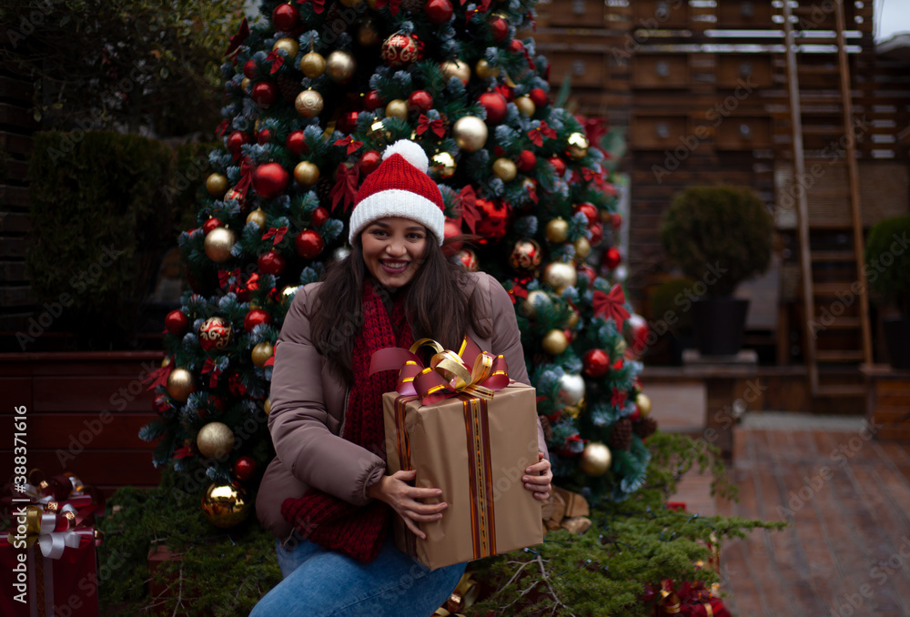 Young pretty woman holds the gift outdoor at the background decorated christmas tree with lights garland on New Year's Eve holidays. Kid dressed warm jacket, red knitted hat Young pretty woman holds the gift outdoor at the background decorated christmas tree with lights garland on New Year's Eve holidays. Kid dressed warm jacket, red knitted hat