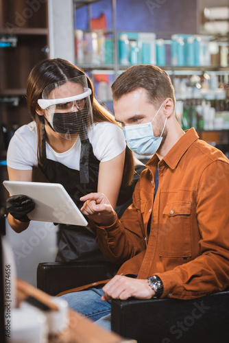 barber in face shield and latex gloves holding digital tablet near client pointing with finger, blurred foreground