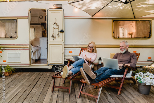 Pleased happy aged couple reading a book and using laptop while relaxing on the deck chairs near the camper van