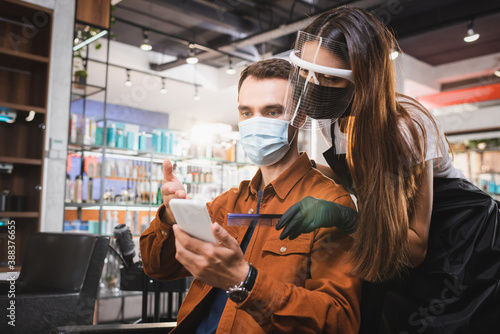 Barber in face shield holding comb near client in medical mask pointing with hand at smartphone