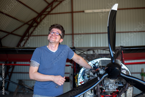 A man repairing airplane engine on small aircraft. Satisfied man holding instruments and fixing detail in the plane engine in hangar. Small aviation. Red aircraft with propeller and cone.