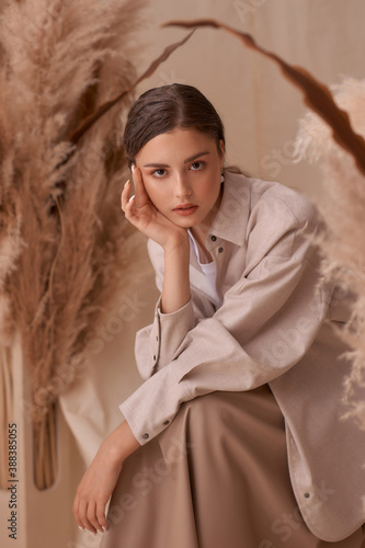 Fall fashion portrait. Brunette girl with pony tail and natural makeup. Elegant woman posing between ears of rye at textile background. Stylish female model in brown skirt anf beige jacket