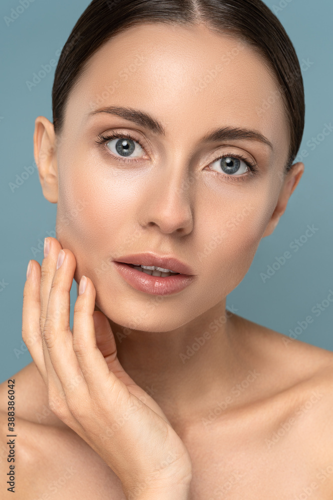 Studio portrait of young woman with natural makeup, combed hair, touching her well-groomed pure skin on face, looking at camera over blue background. Beauty, facelift, anti-aging cosmetic procedure. Studio portrait of young woman with natural makeup, combed hair, touching her well-groomed pure skin on face, looking at camera over blue background. Beauty, facelift, anti-aging cosmetic procedure.