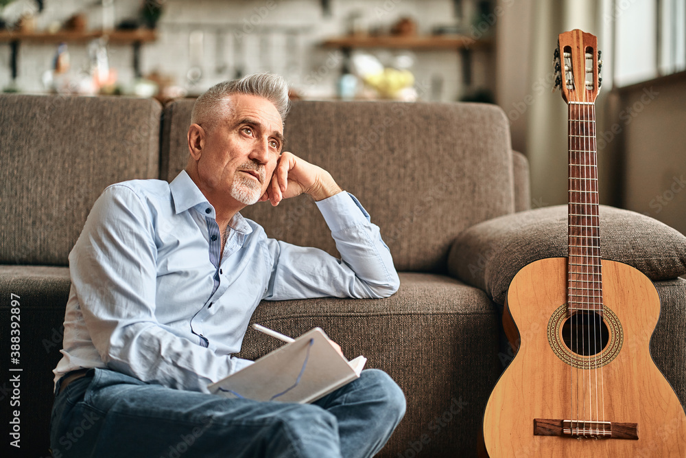 Thoughtful adult gray-haired man writing song with guitar in the living room