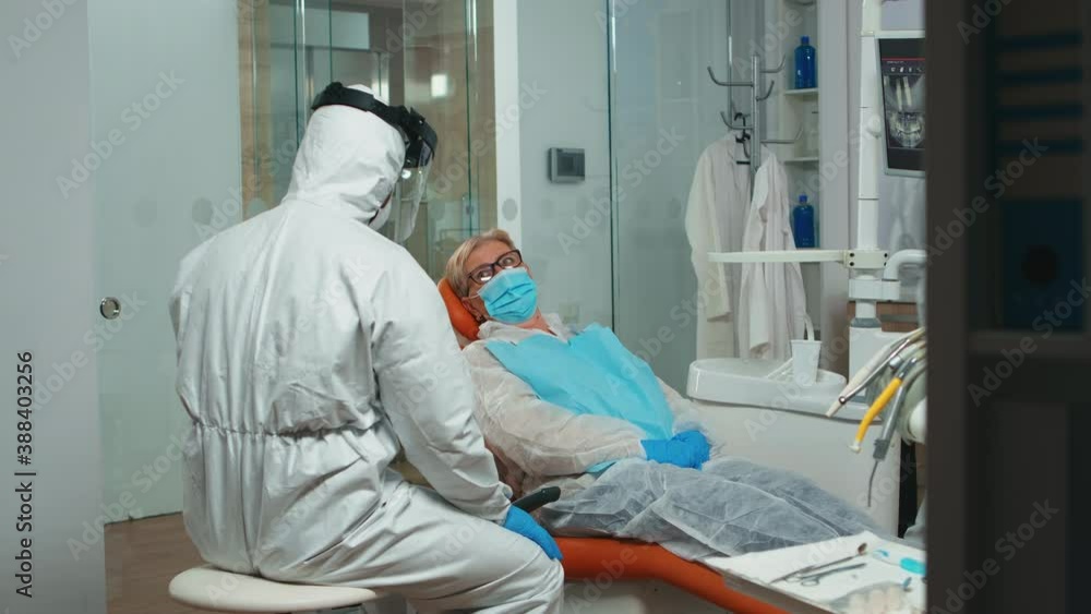 Dentist doctor in coverall showing to senior patient x-ray on stomatological monitor during coronavirus. Concept of new normal dentist visit in coronavirus outbreak wearing protective suit and face Dentist doctor in coverall showing to senior patient x-ray on stomatological monitor during coronavirus. Concept of new normal dentist visit in coronavirus outbreak wearing protective suit and face