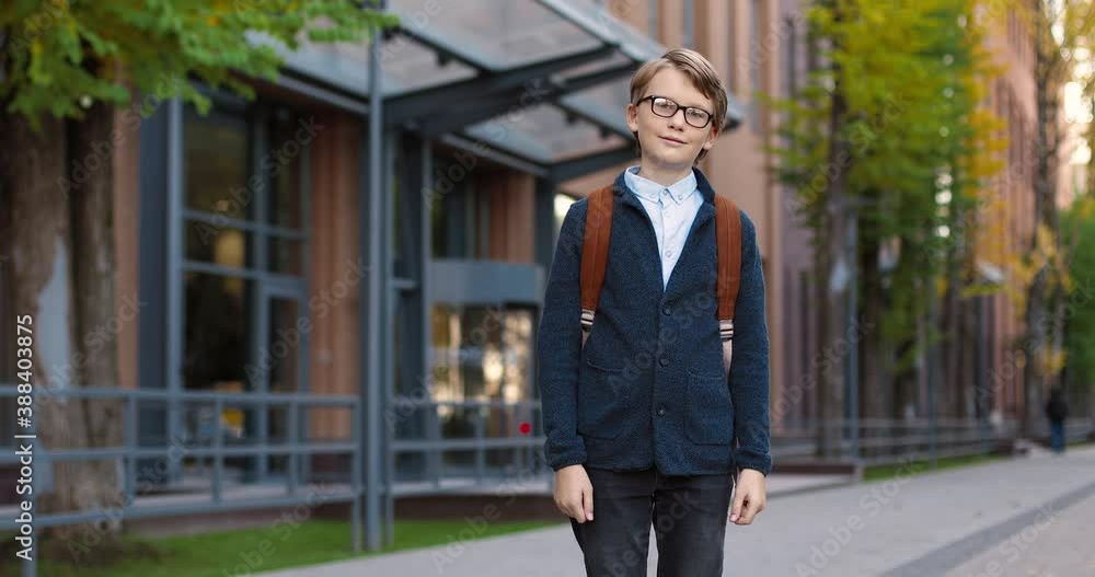 Portrait of joyful Caucasian boy pupil in glasses in good mood standing outdoor near school. Happy male student with backpack smiling to camera on street in town. Education concept Portrait of joyful Caucasian boy pupil in glasses in good mood standing outdoor near school. Happy male student with backpack smiling to camera on street in town. Education concept