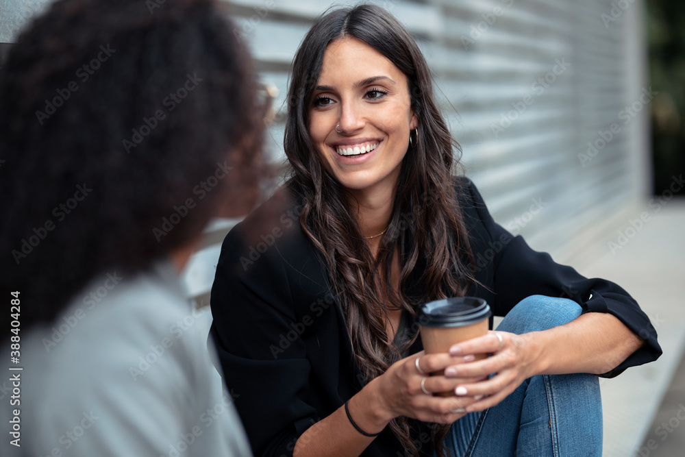 Two beautiful businesswomen talking while drinking coffee sitting on a bench on the street. Two beautiful businesswomen talking while drinking coffee sitting on a bench on the street.