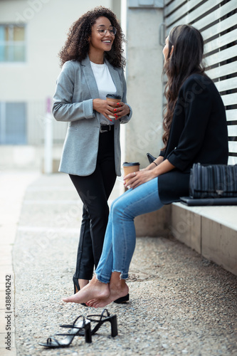 Two beautiful businesswomen talking while drinking coffee sitting on a bench on the street. © nenetus