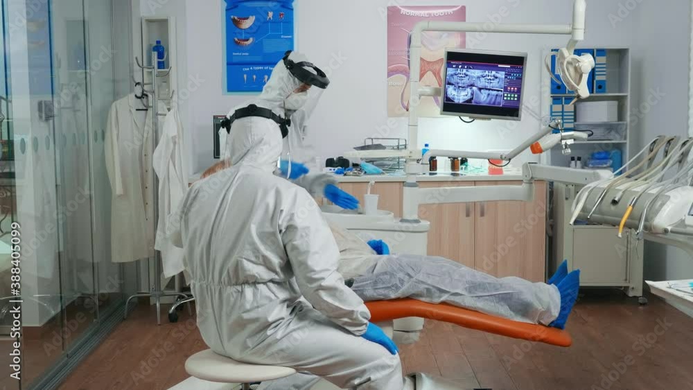 Dental specialist with coverall speaking with senior patient before examination during pandemic covid-19. Assistant and orthodontic doctor wearing protection suit, face shield, mask and gloves. Dental specialist with coverall speaking with senior patient before examination during pandemic covid-19. Assistant and orthodontic doctor wearing protection suit, face shield, mask and gloves.