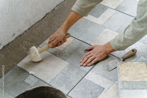 Close up on hands of unknown senior man craftsman using hammer to adjust and lay ceramic tiles on the balcony or terrace over adhesive cement in day - construction industry concept copy space