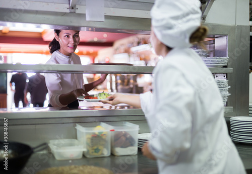 Smiling waitress taking cooked ordered meals in restaurant kitchen for serving guests