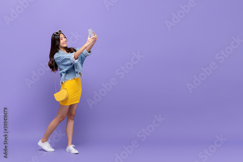 Full length portrait of smiling young pretty Asian woman taking selfie with smartphone in isolated studio purple background with copy space