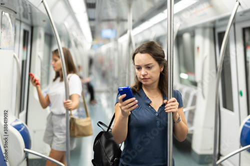 Young focused woman standing in subway car holding on handrails, browsing in her smartphone..