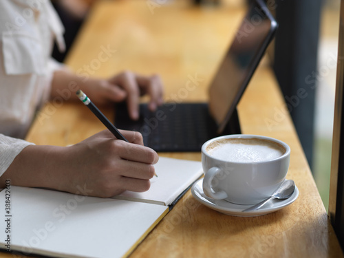 Female hand writing on blank notebook while working with tablet on wooden bar