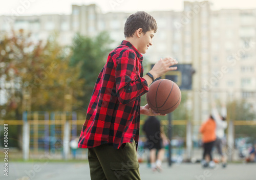Cute young boy plays basketball on street playground. Teenager in red check flannel shirt with orange basketball ball outside. Hobby, active lifestyle, sport activity for kids. © Natali
