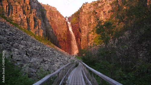 wide shot from afar of majestic Njupeskärs, Sweden's highest waterfall lit by midsummer morning golden sun, guided by wooden passageway in  Fulufjällets Nationalpark
 photo