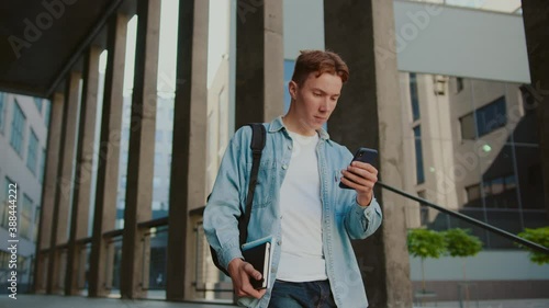 Close up student young handsome man with red hair walking down the stairs near university or office building. Use smartphone in casual jeans. Look around. Slow motion