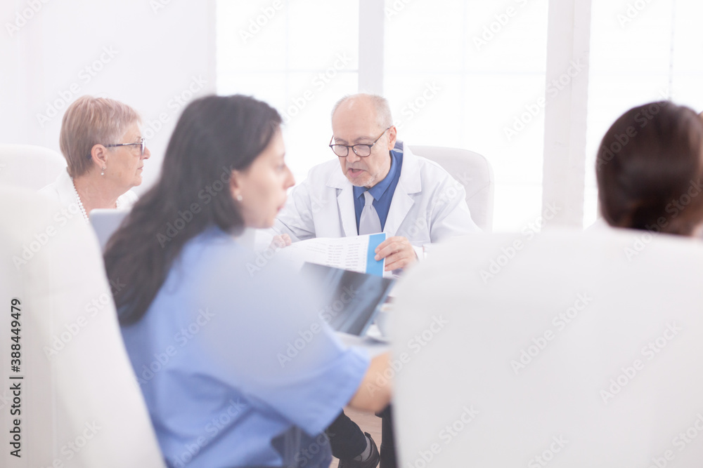 Mature doctor reading health raport during meeting with coworkes in hospital conference room. Clinic expert therapist talking with colleagues about disease, medicine professional Mature doctor reading health raport during meeting with coworkes in hospital conference room. Clinic expert therapist talking with colleagues about disease, medicine professional