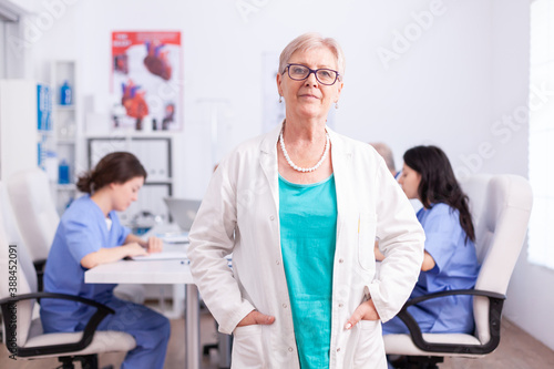 Portrait of senior doctor smiling at camera in hospital conference room with medical staff in the background.