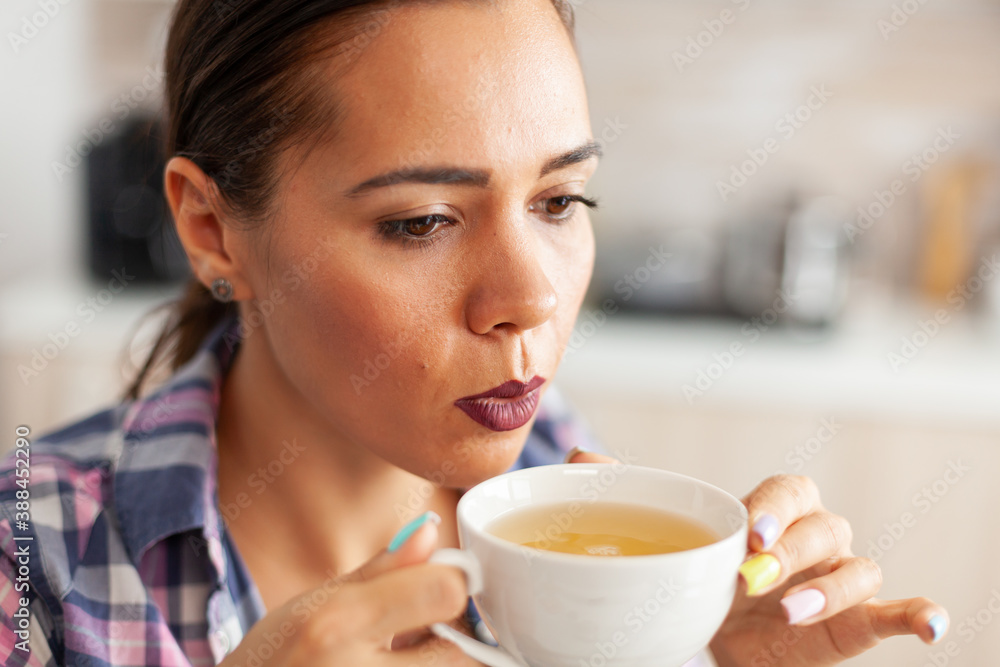 Close up of woman in kitchen trying to drink hot green tea with aromatic herbs. Pretty lady sitting in the kitchen in the morning during breakfast time relaxing with tasty natural herbal tea.