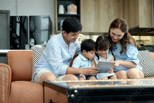 Asian family young parents sit on couch with daughter and son have fun using tablets together to watch funny videos in living room at home. Activities together and relationships in family concept.