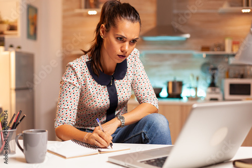 Businesswoman writing notes while working late on computer in home kitchen. Employee using modern technology at midnight doing overtime for job, business, busy, career, network, lifestyle ,wireless. photo