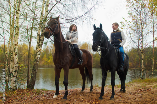 Cute young couple on horsebacks in the autumn forest by lake. Riders in autumn Park in inclement cloudy weather with light rain. Concept of outdoor riding, sports and recreation. Copy space