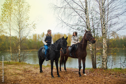 Cute young couple on horsebacks in the autumn forest by lake. Riders in autumn Park in inclement cloudy weather with light rain. Concept of outdoor riding, sports and recreation. Copy space © Alex Vog