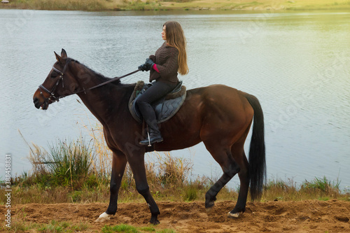 Cute young woman on horseback in autumn forest by lake. Rider female drives her horse in Park in inclement cloudy weather with rain. Concept of outdoor riding, sports and recreation. Copy space