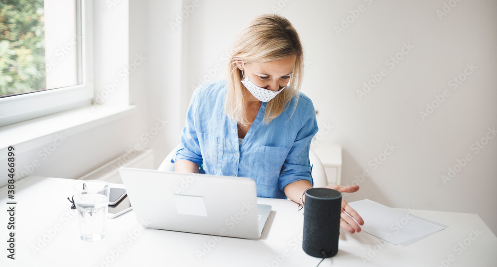 beautiful young and blond business woman wearing nose mouth mask talking to smart speaker in office during covid 19 crisis beautiful young and blond business woman wearing nose mouth mask talking to smart speaker in office during covid 19 crisis