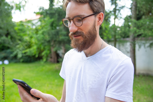 Young bearded man standing outdoor with a mobile phone in his hand. Receiving a message on a cell phone, smartphone.