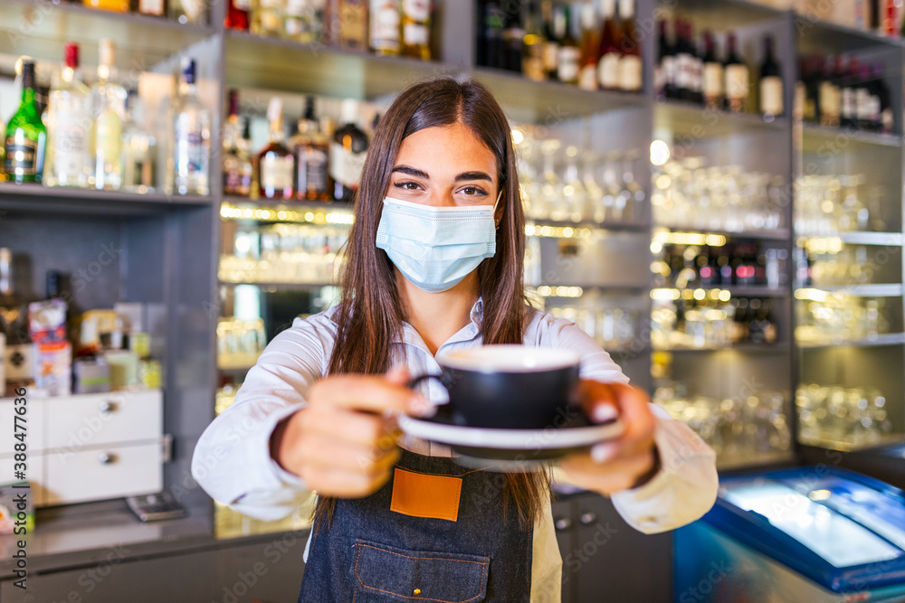 Beautiful female barista is holding a cup with hot coffee, looking at camera and wearing protective face mask while standing near the bar counter in cafe