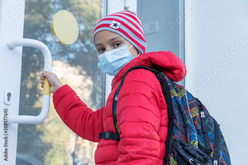 schoolboy in medical face mask looking into camera and pulling door before entering school. New reality, restrictions to prevent covid pandemic. Healthcare concept photo