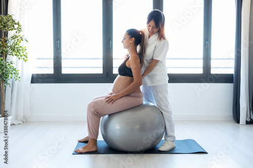 Beautiful young physiotherapist helping to beautiful pregnant woman for doing pilates exercises with ball preparing for childbirth at home.