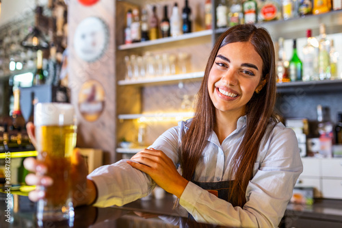 Beautiful smiling female Bartender serving a draft beer at the bar counter , shelves full of bottles with alcohol on the background
