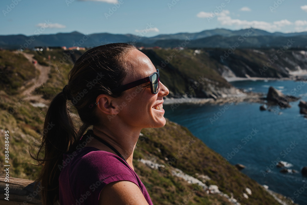 Side portrait of white woman with sunglasses looking at the sea from the top of a cliff.