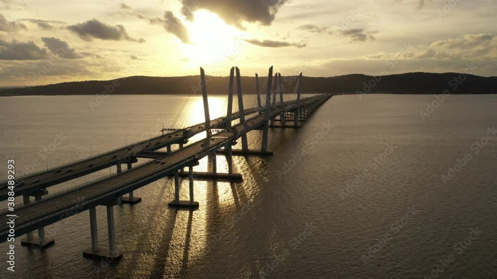 An aerial shot along the north side of the Mario M. Cuomo Bridge during a golden sunset. The drone camera dolly out over the Hudson River flying away from South Nyack, NY in the background. An aerial shot along the north side of the Mario M. Cuomo Bridge during a golden sunset. The drone camera dolly out over the Hudson River flying away from South Nyack, NY in the background.