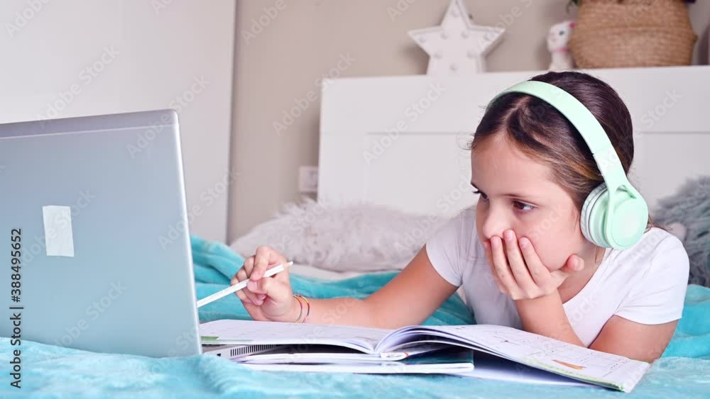 Online lessons. A little girl, a schoolgirl in headphones, with textbooks, makes a lesson on the computer. New version of homeschooling during the quarantine period, covid 19. 4k footage Online lessons. A little girl, a schoolgirl in headphones, with textbooks, makes a lesson on the computer. New version of homeschooling during the quarantine period, covid 19. 4k footage