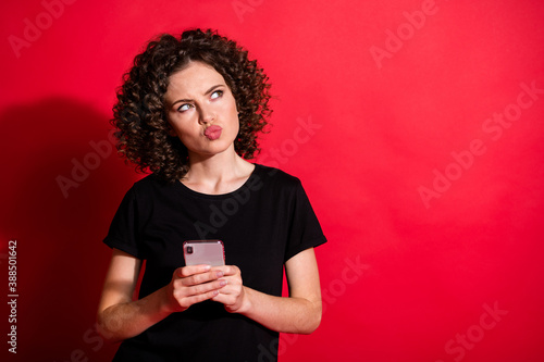 Photo of thoughtful curly woman wear casual black outfit looking empty space phone hands arms lips pouted isolated red color background © deagreez