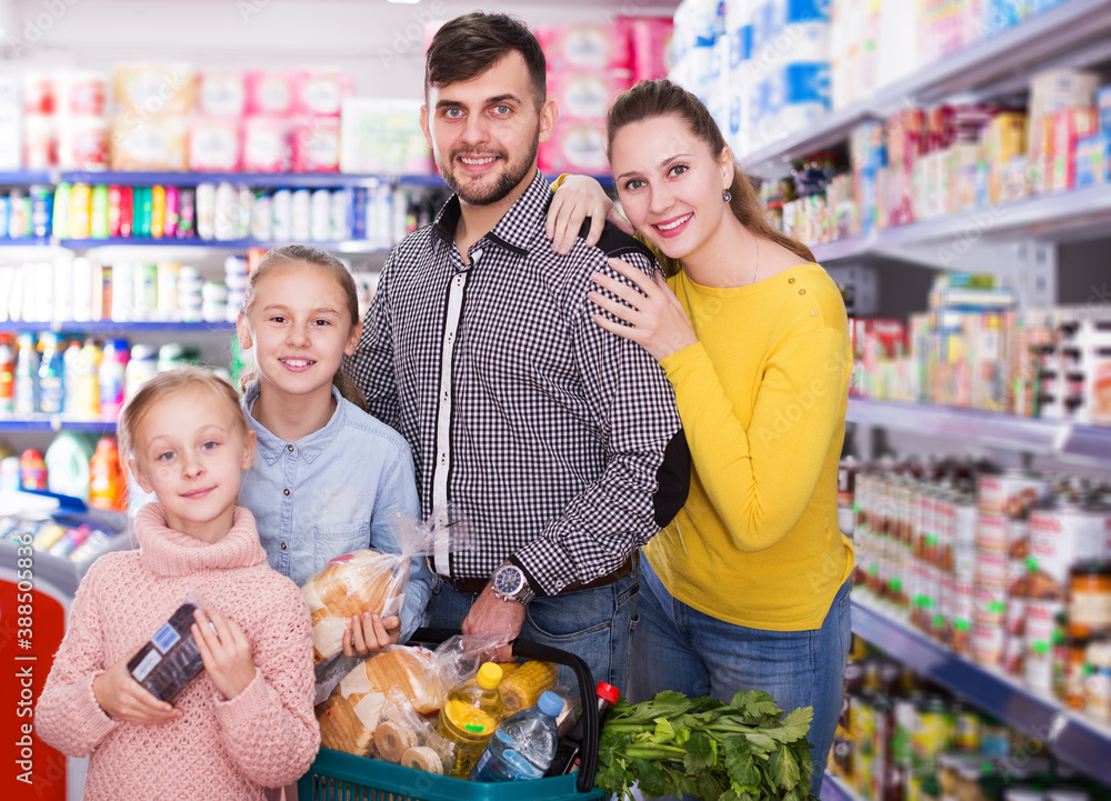 smiling young parents with two little girls with purchases during family shopping in grocery store