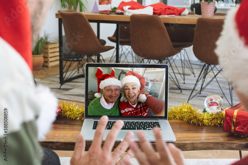 Rear view of man and son in santa hats waving while having a videocall with senior couple in santa h