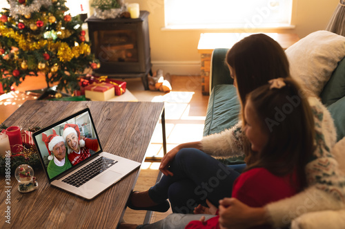 Woman and daughter sitting on couch having a videocall with senior couple in santa hats smiling on l